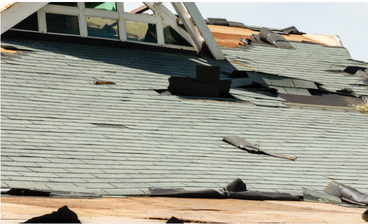 Damaged asphalt shingle roof with missing sections, debris, and a window in the background.