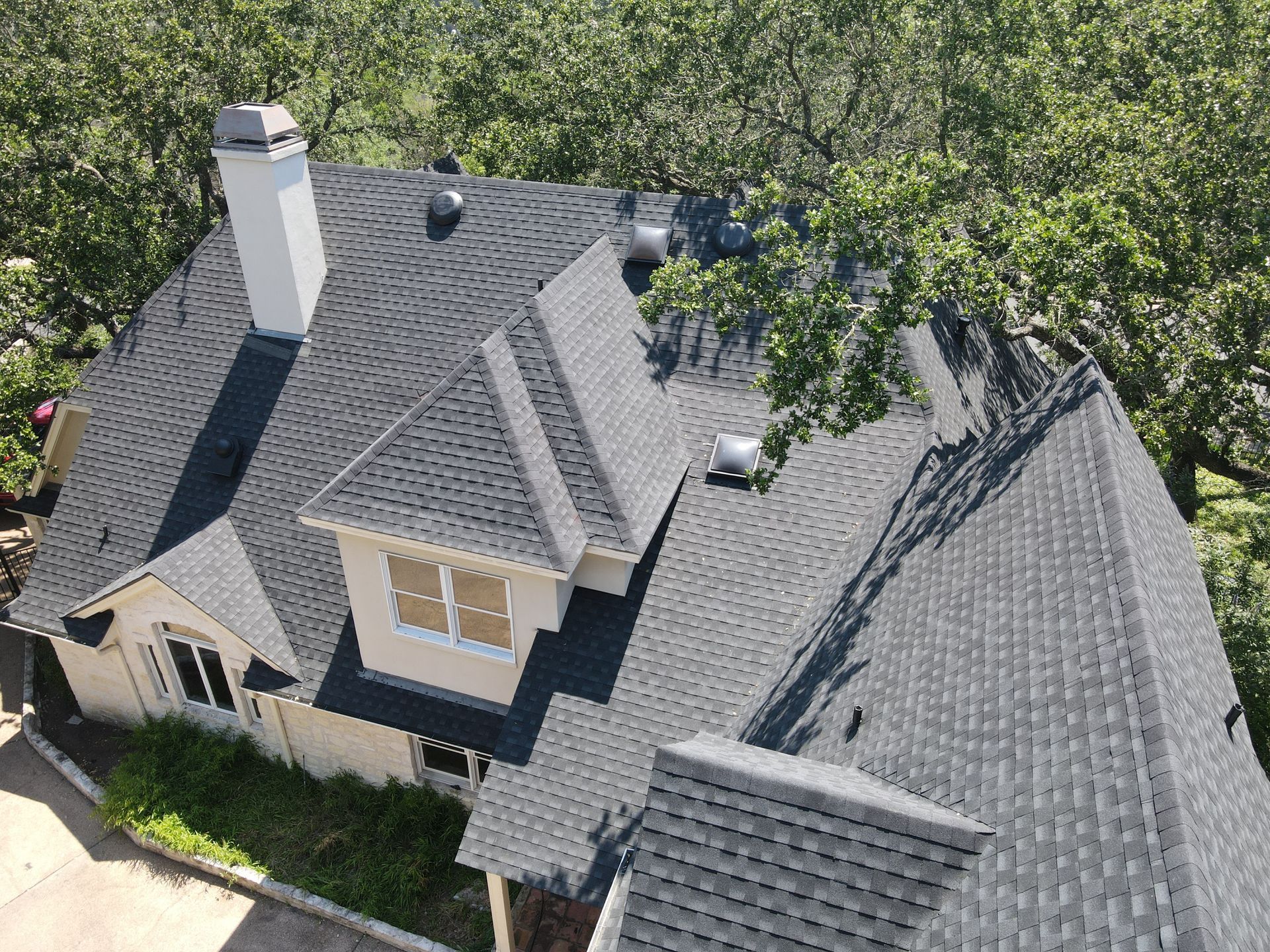 Overhead view of a house with a dark gray shingle roof, chimney, and surrounded by trees.