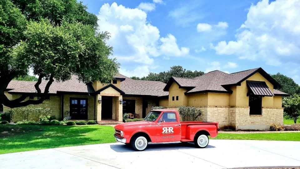 Red vintage pickup truck parked in front of a yellow and stone house on a sunny day.