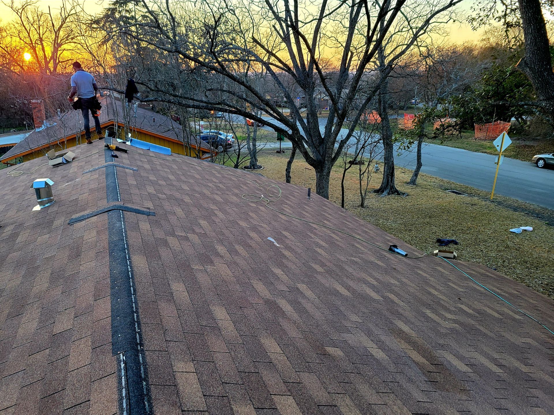 Roofer on a brown shingled roof, near bare trees, setting sun in the background, and a residential street.