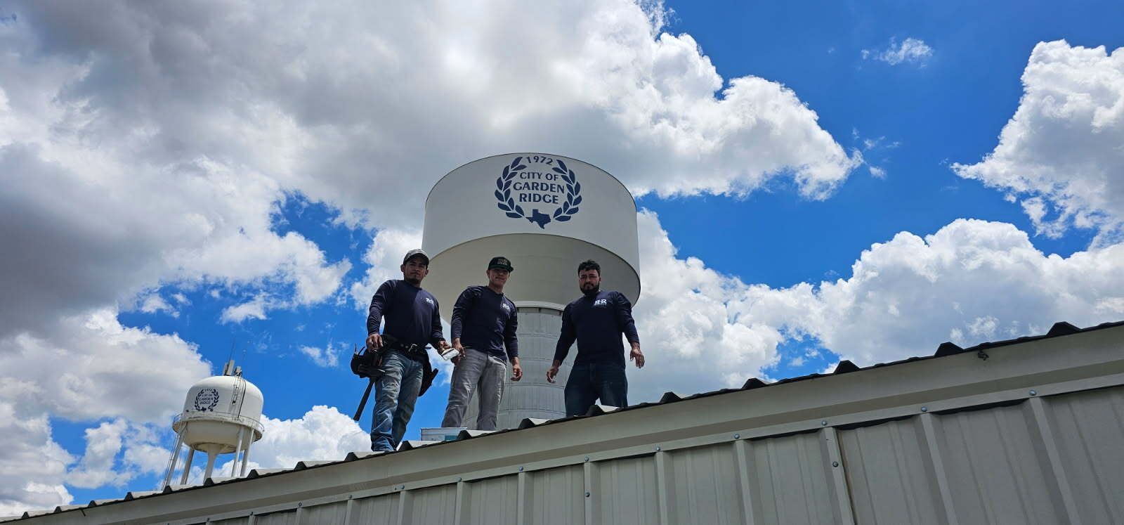 Three men on a roof near a water tower on a bright, cloudy day.