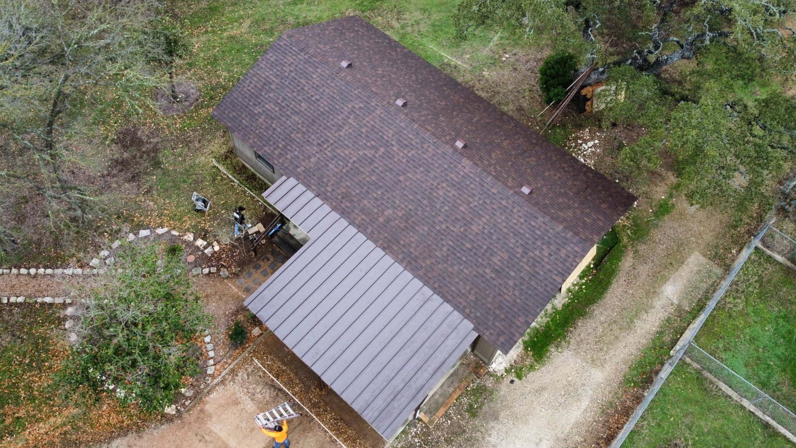 Aerial view of a house with a brown roof and a grey metal awning, surrounded by trees and a gravel driveway.