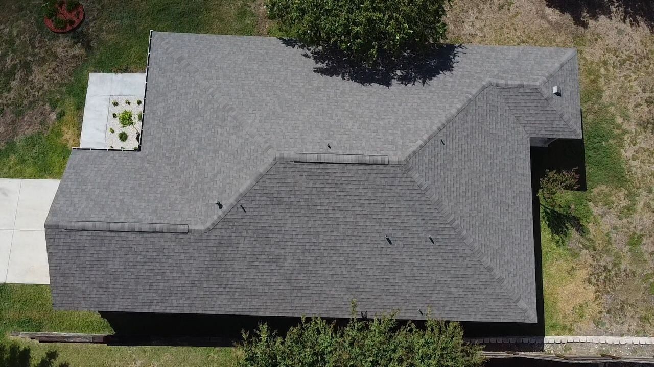 Overhead view of a gray shingled roof of a house, next to a driveway and grassy yard.