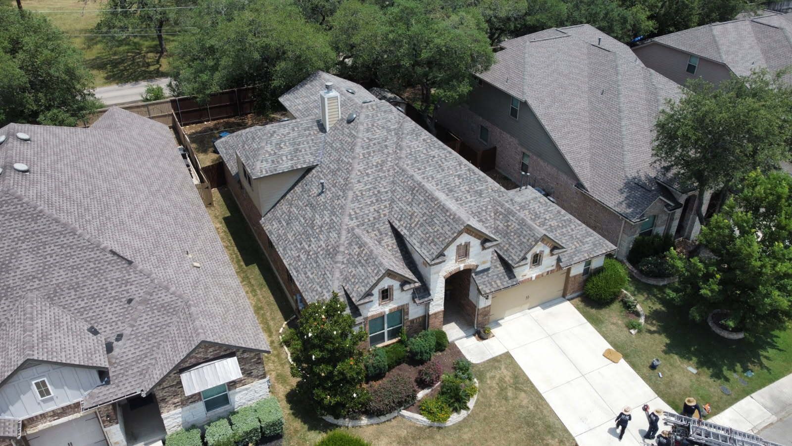 Aerial view of a residential home with a gray roof, concrete driveway, and surrounding trees and homes.