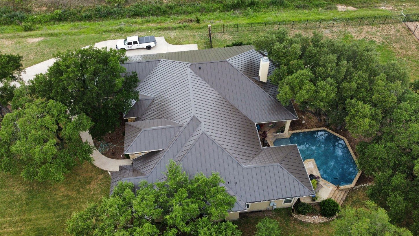 Aerial view of a house with a dark roof, a pool, and surrounded by trees and grass.