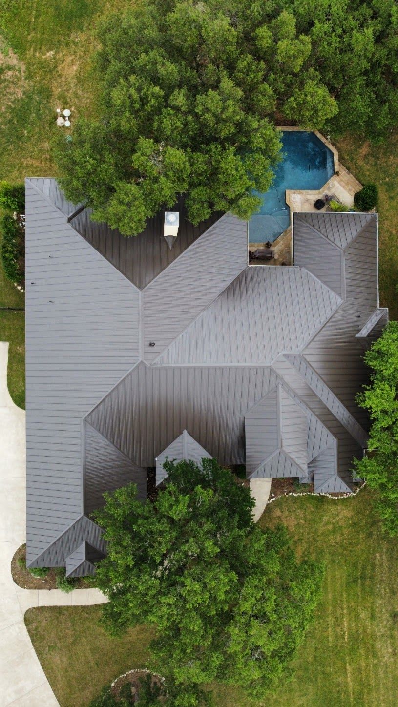 Overhead view of a house with a dark gray roof, pool, and surrounding trees and grass.