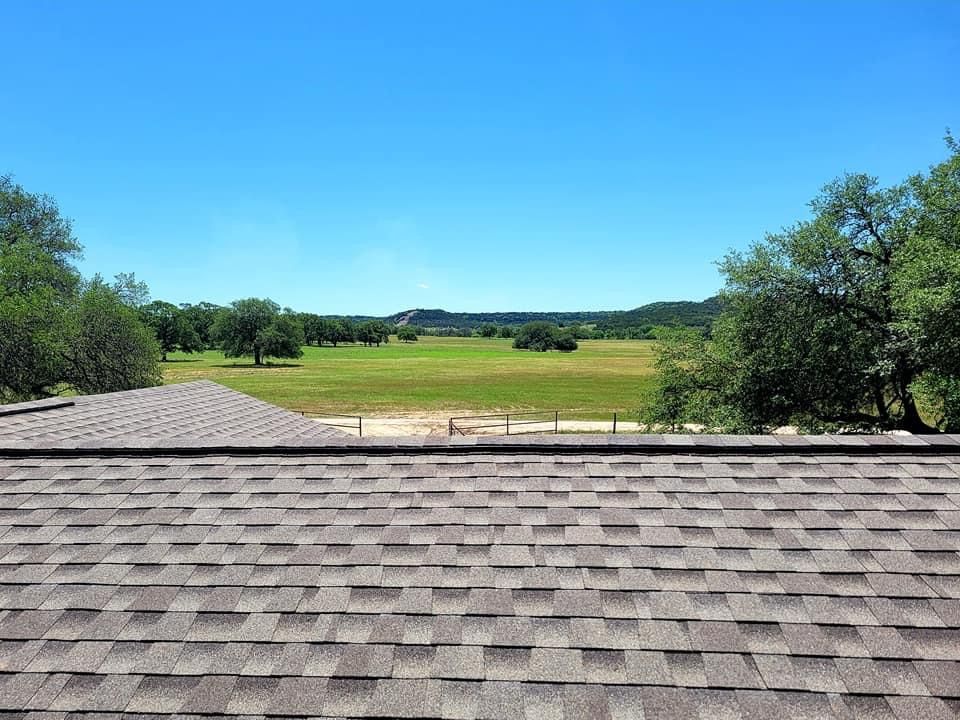 Brown shingled roof overlooks green field, trees, and clear blue sky on a sunny day.