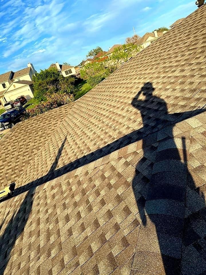 Shadow of person on a brown shingled roof; sunny day, blue sky, houses in background.