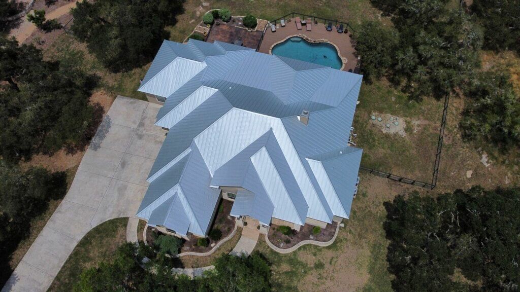 Aerial view of a house with a gray metal roof, driveway, and a pool surrounded by trees.