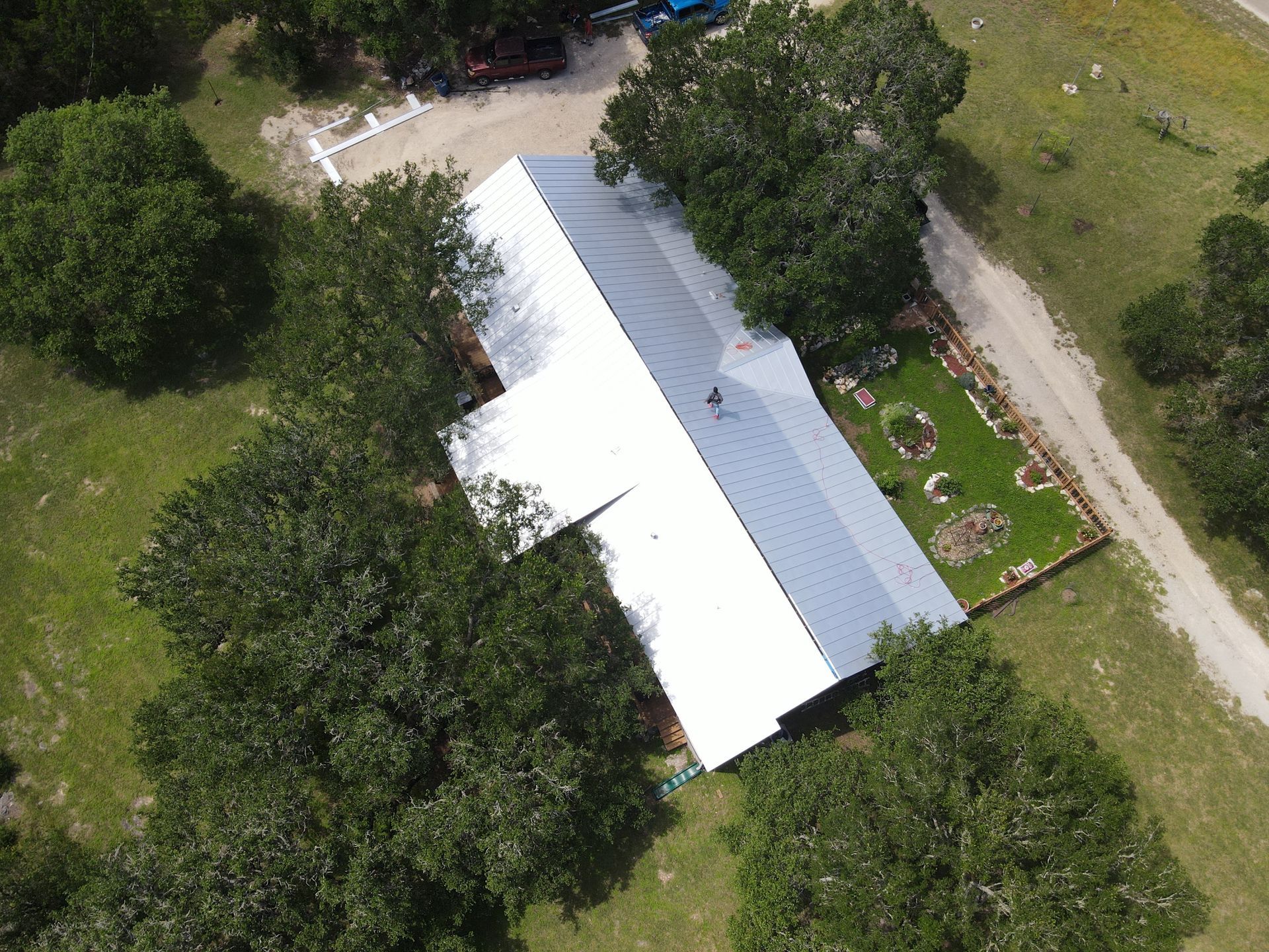 Aerial view of a house with a white metal roof surrounded by green trees and a small garden.