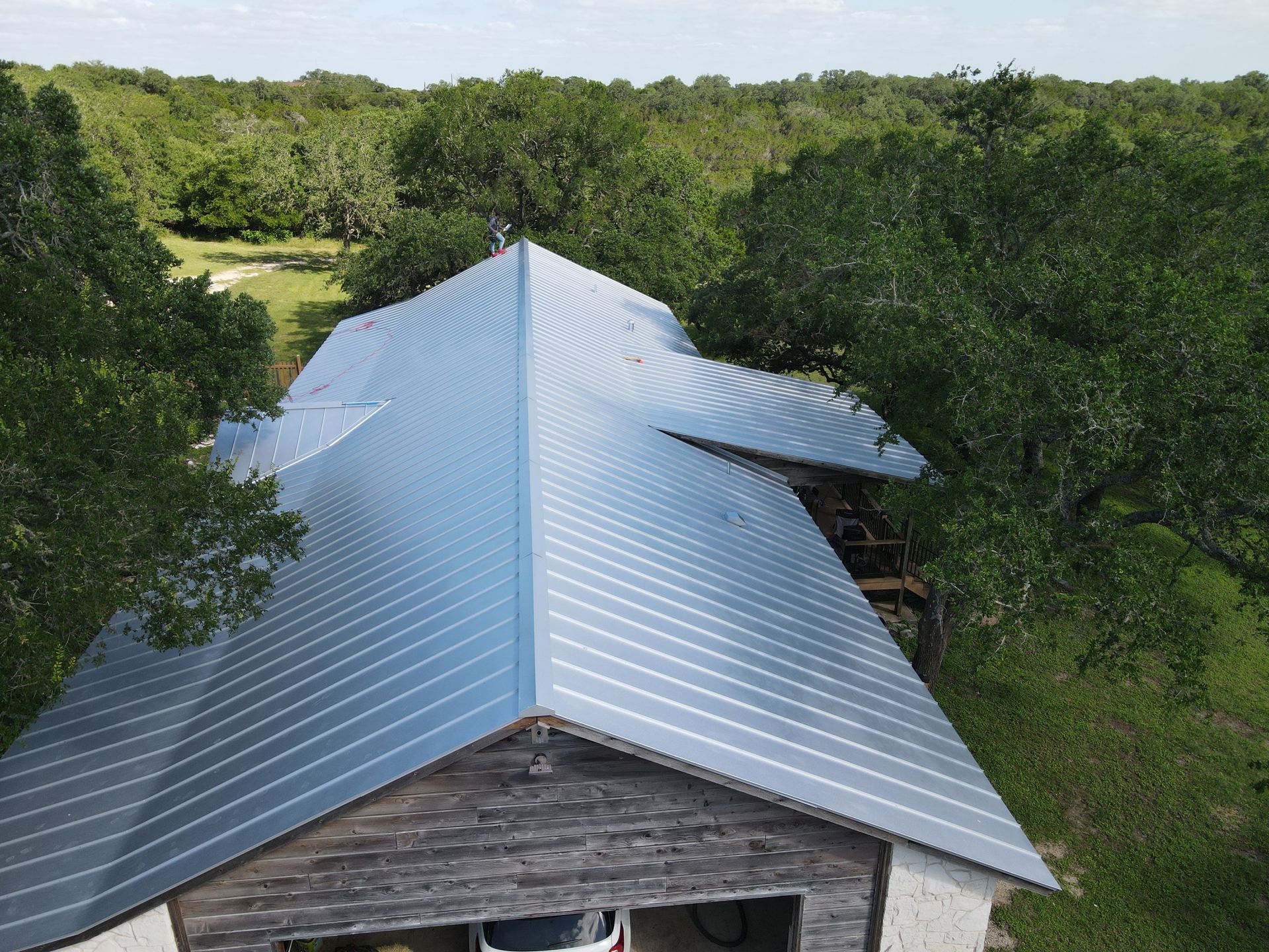 An overhead view of a gray metal roof on a rustic building surrounded by green trees.