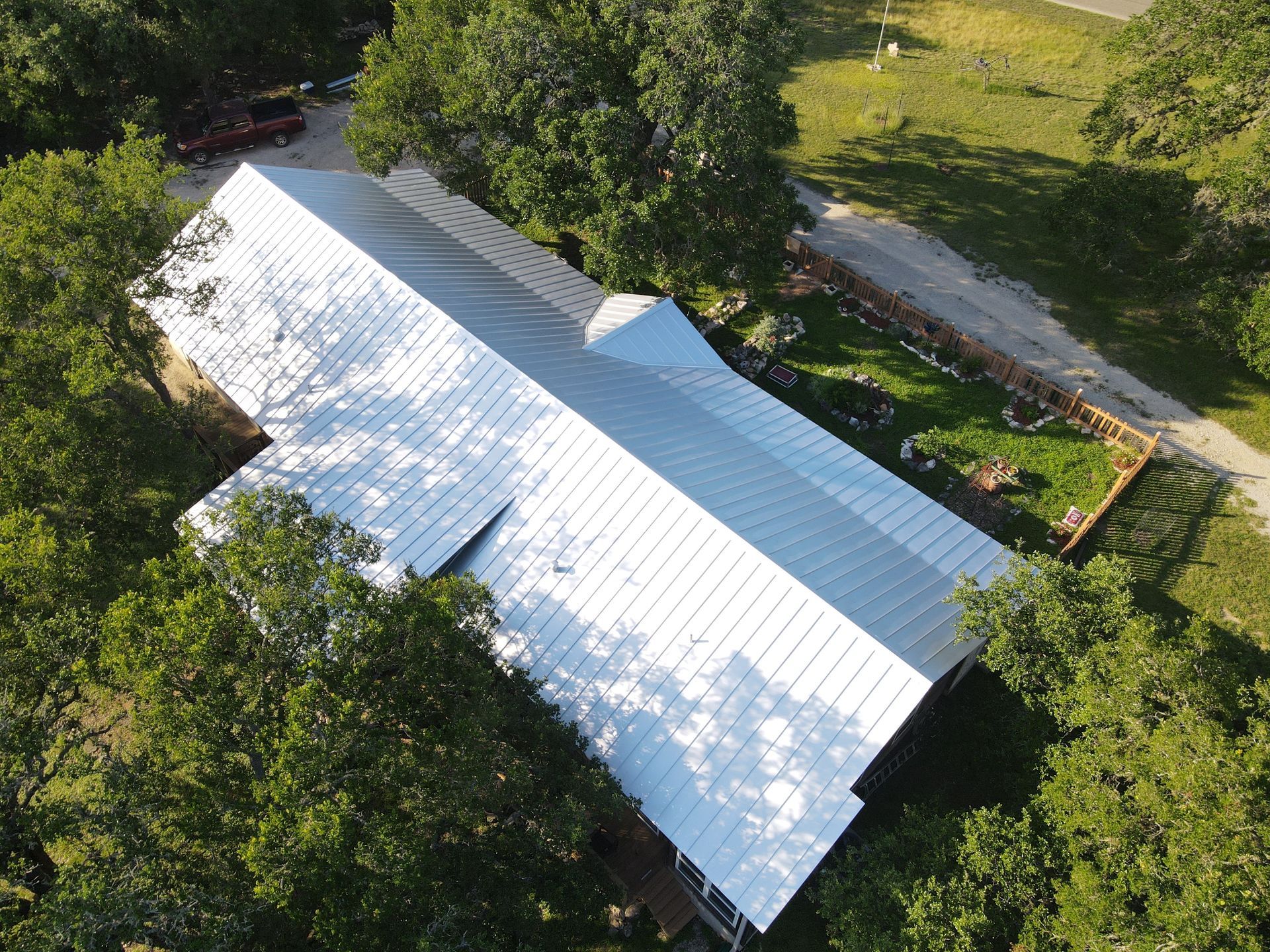 Aerial view of a long building with a shiny metal roof, surrounded by green trees and grass.