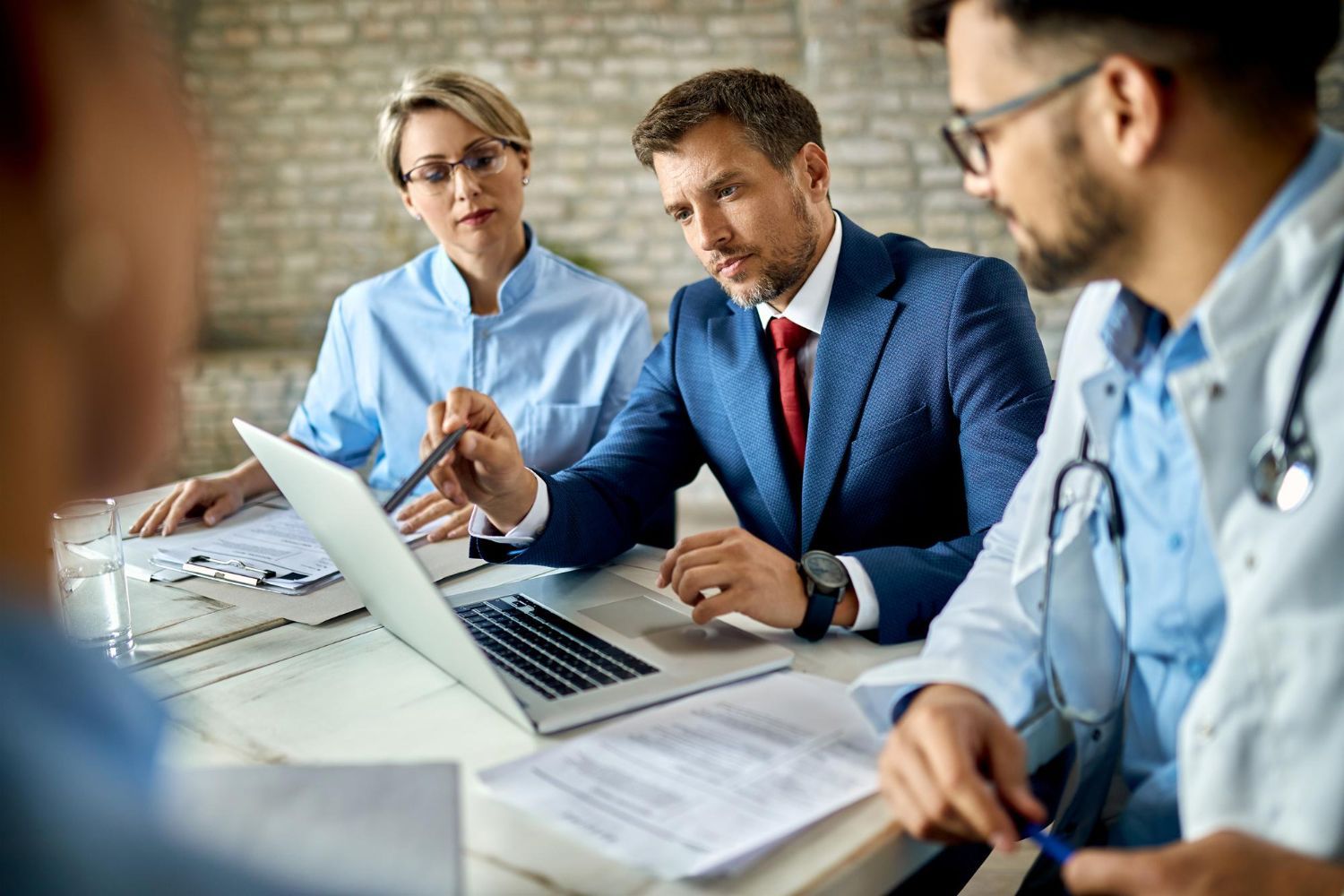 A group of doctors and business people are sitting around a table looking at a laptop.