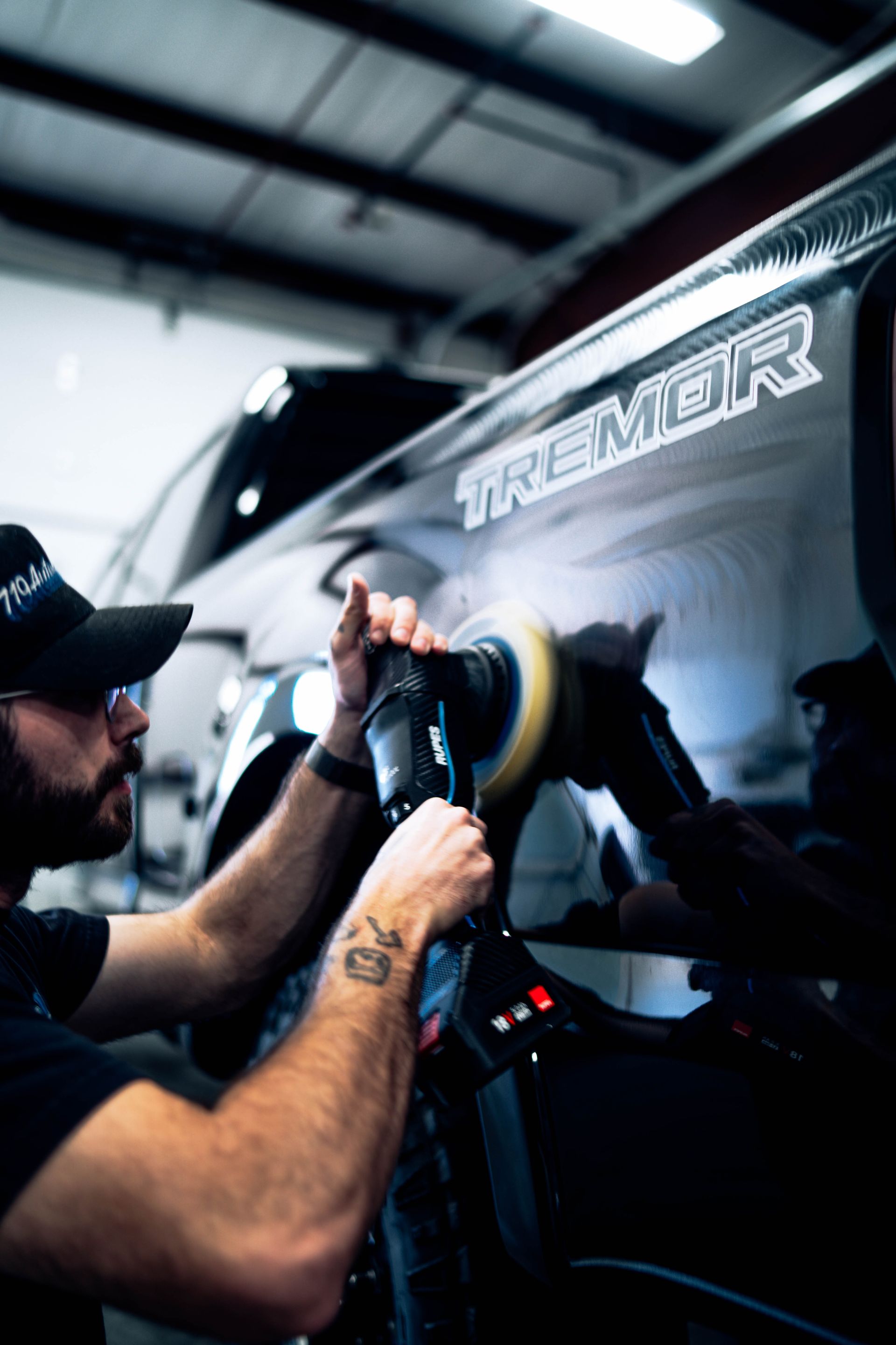 Mechanic polishing a black car in a garage, with a buffing wheel and reflective body panel.