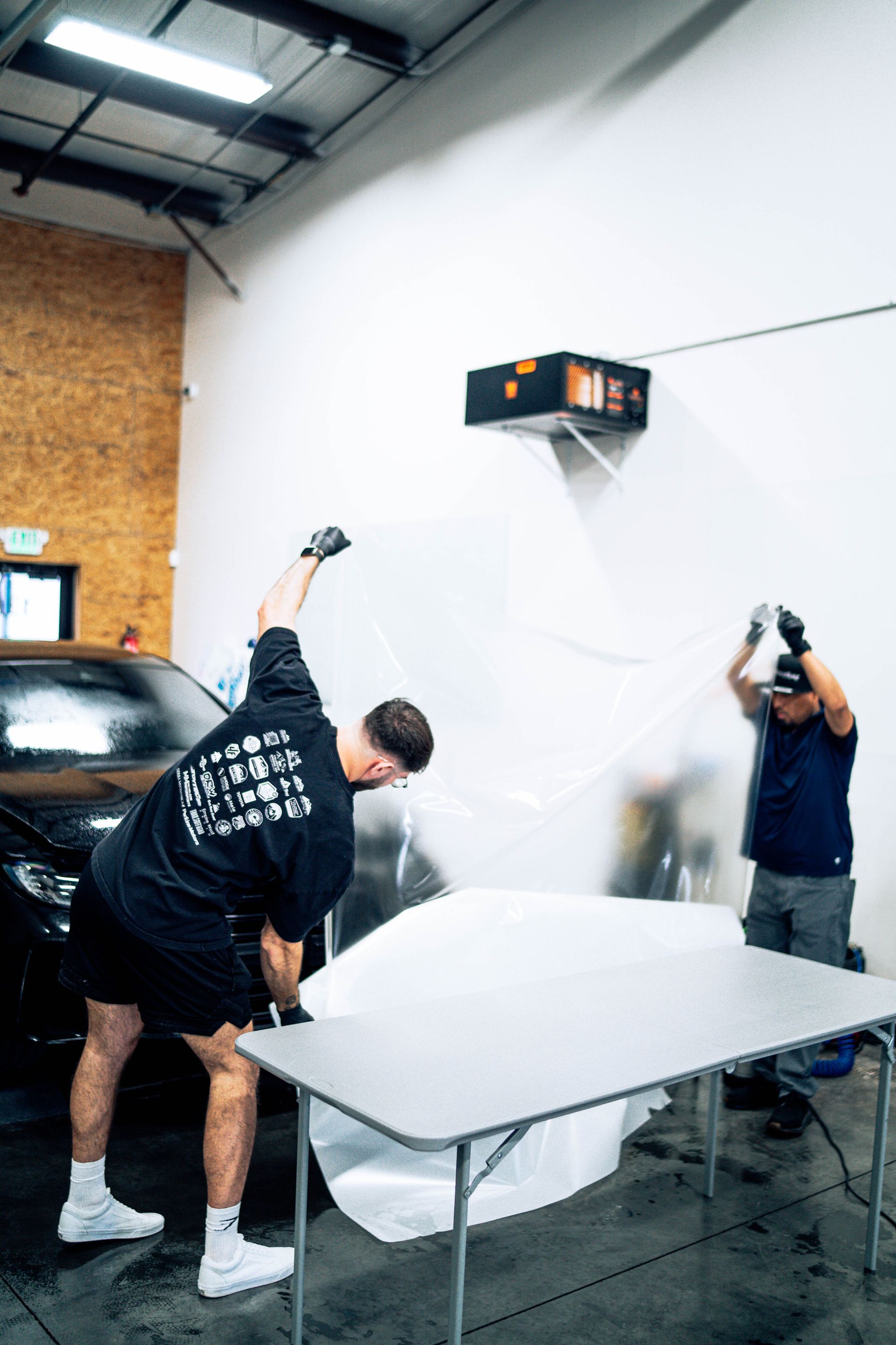 Two people spraying a white tabletop on sawhorses in a workshop with a car and brick wall.