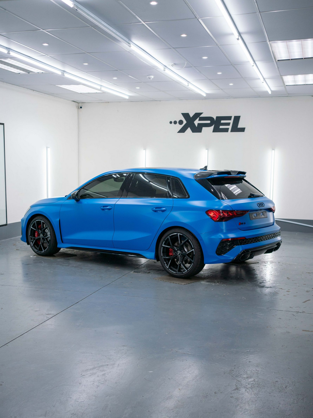 A matte blue Audi RS 3 hatchback parked inside a professional automotive garage with XPEL branding on the white wall.