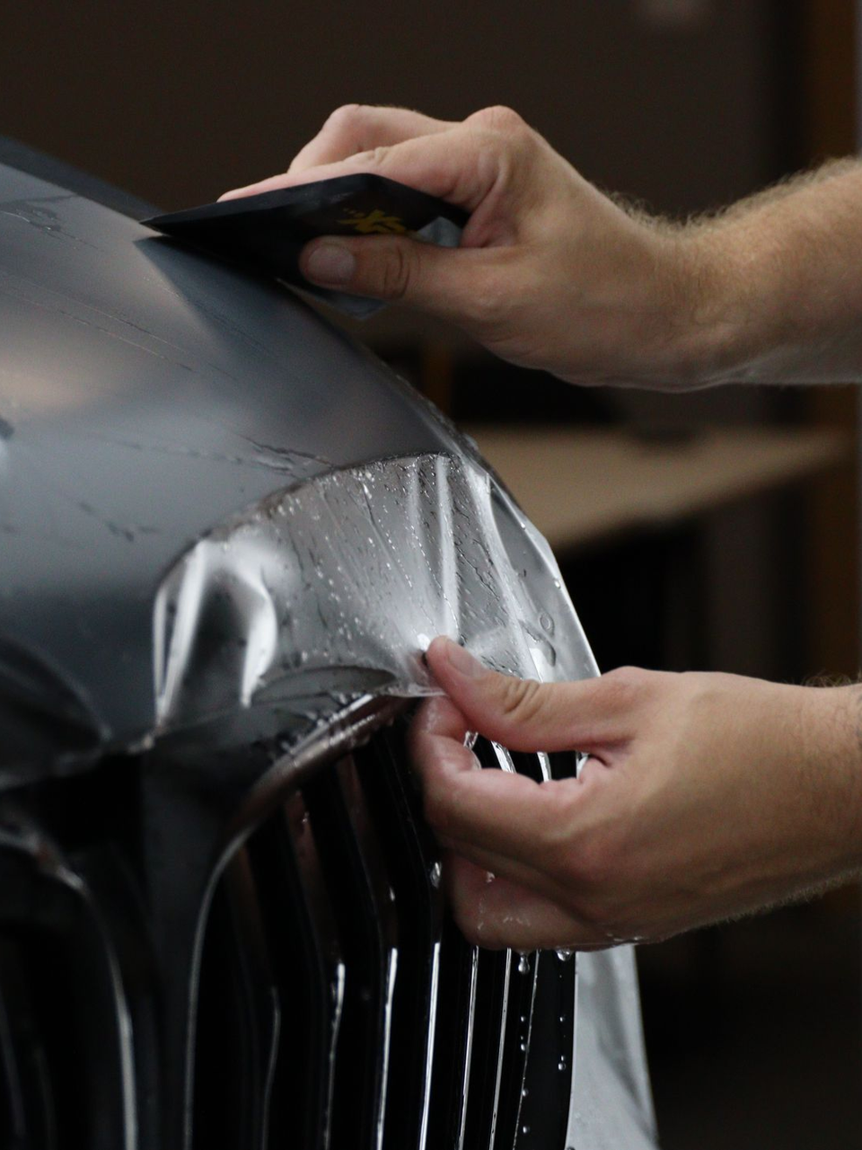 A person uses a squeegee to apply a clear protective film to the gray front bumper of a vehicle.