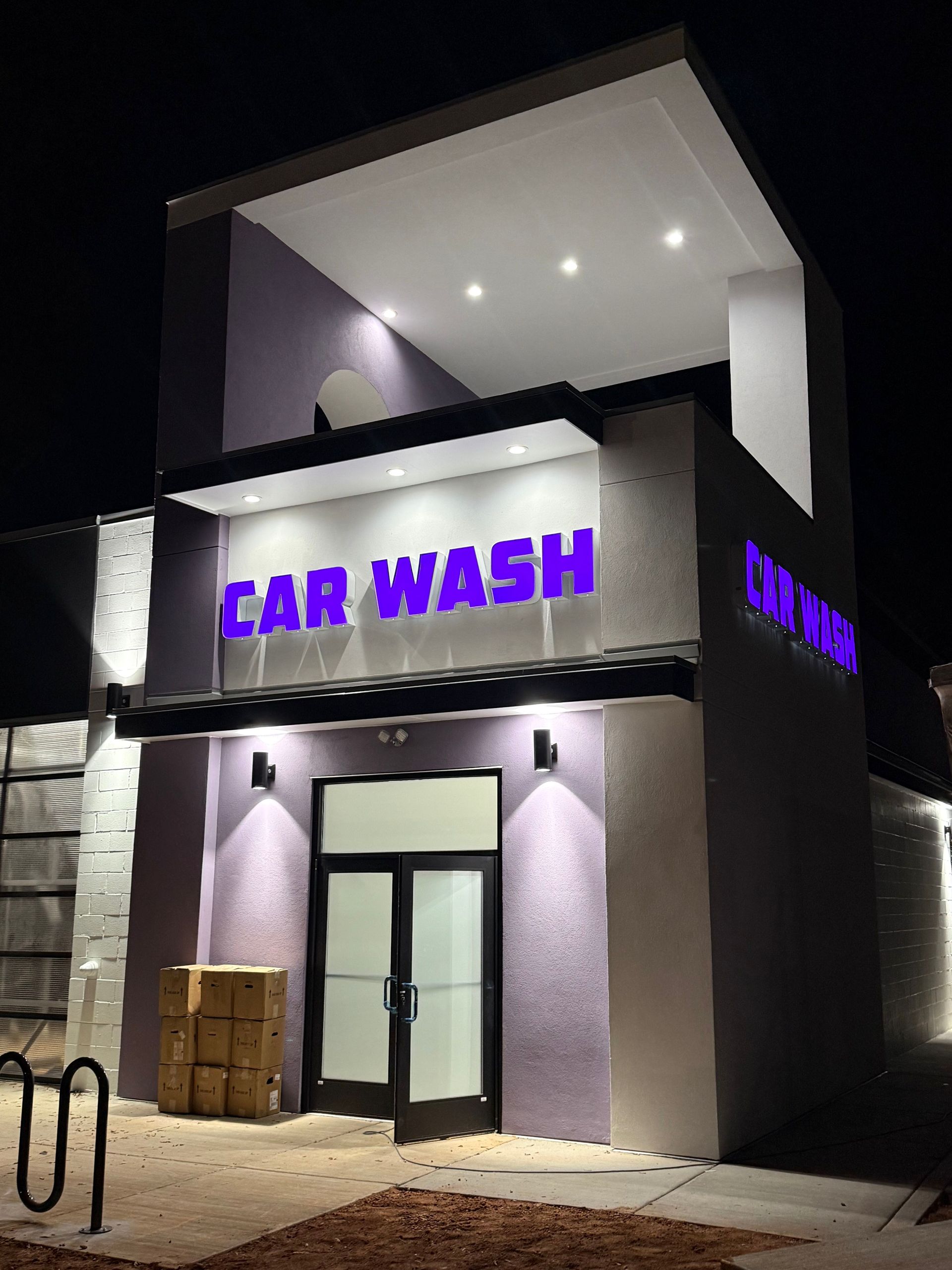 A modern purple and white car wash building at night, featuring illuminated signage and recessed exterior lighting.