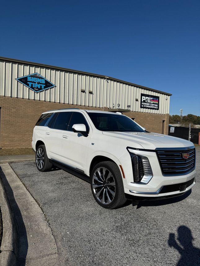 A white Cadillac Escalade parked on a gravel lot in front of a brick building with a 