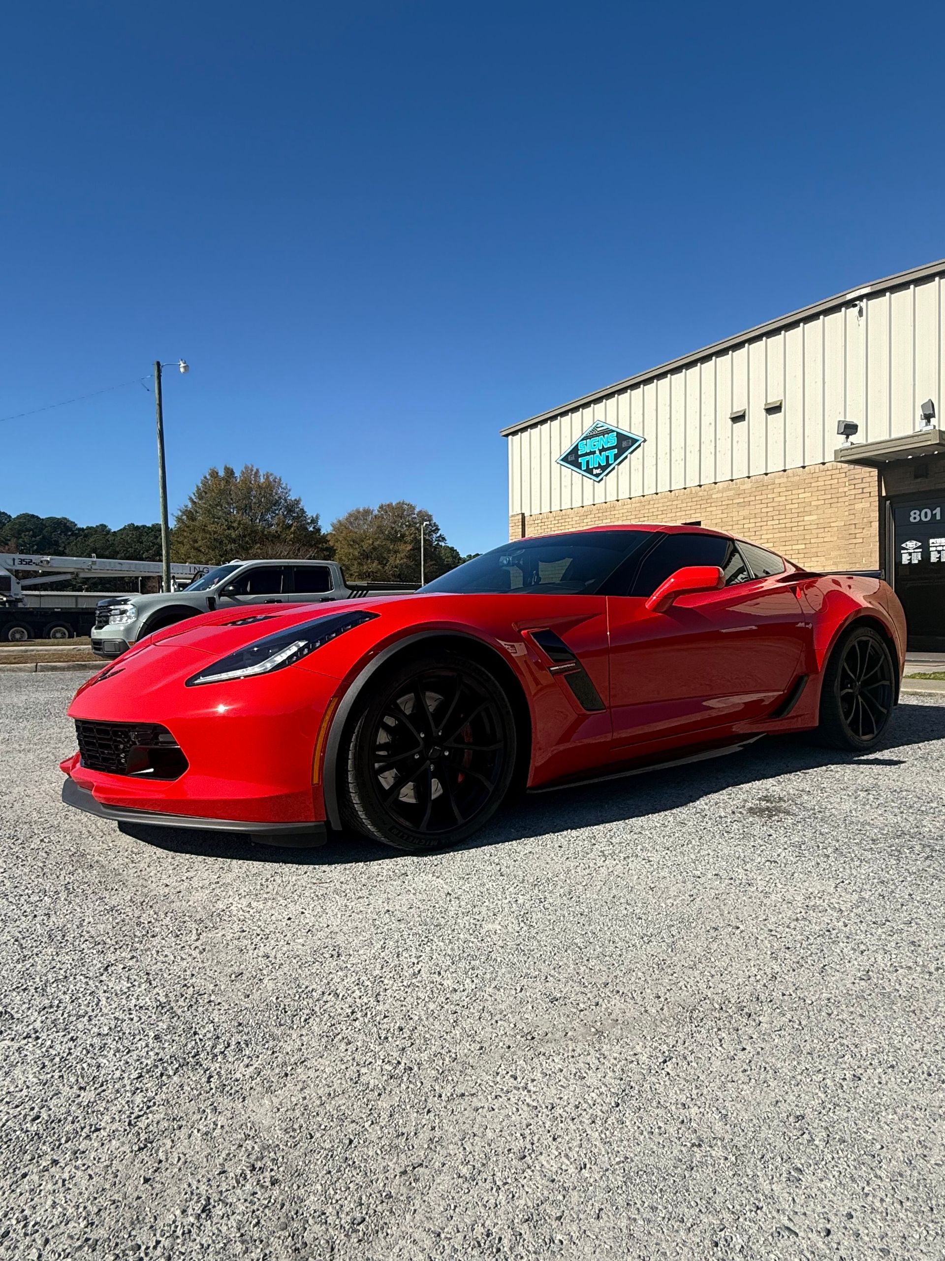 A bright red Chevrolet Corvette parked on a gravel lot in front of a building under a clear blue sky.