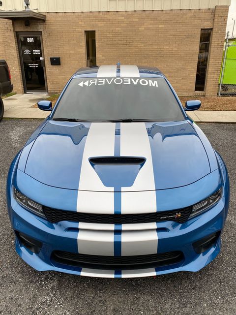 A blue Dodge Charger with silver racing stripes and a "MOVEOVER" windshield banner parked in front of a brick building.