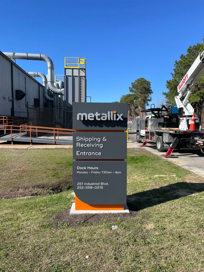 A grey and orange Metallix sign stands on a grassy lawn in front of an industrial facility with a construction lift.