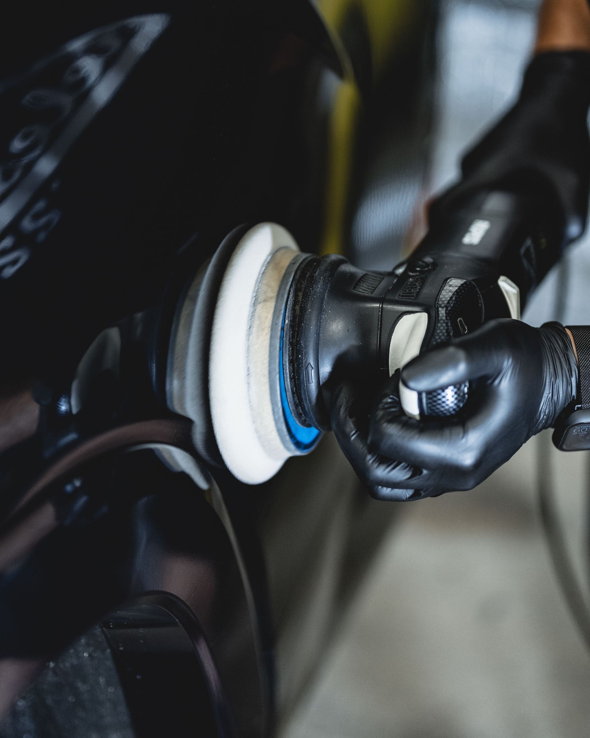 A gloved hand uses a power polisher with a white foam pad to buff the black paint of a car.