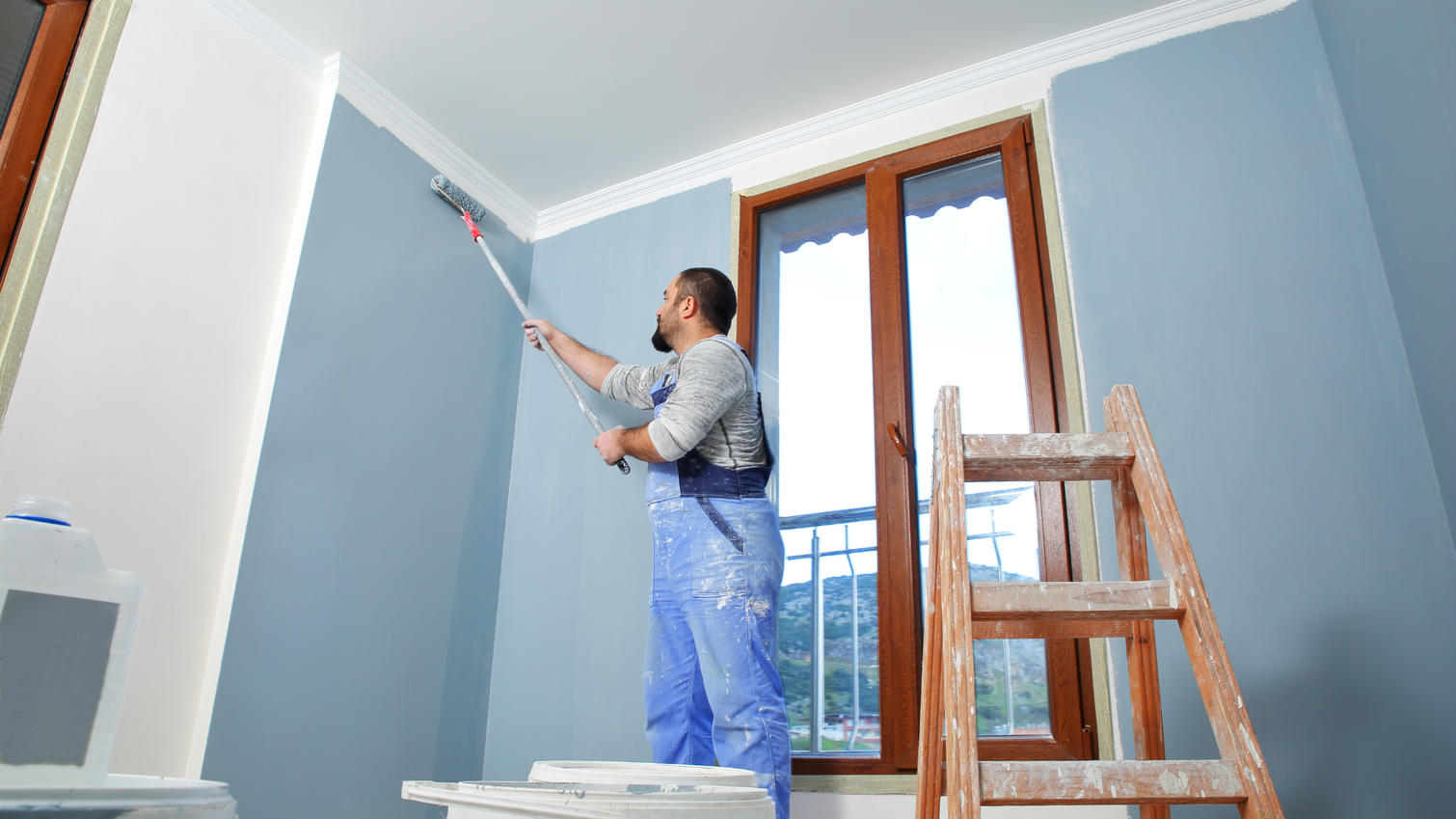 A painter in blue coveralls uses a roller to paint a wall light blue next to a wooden ladder and a window.