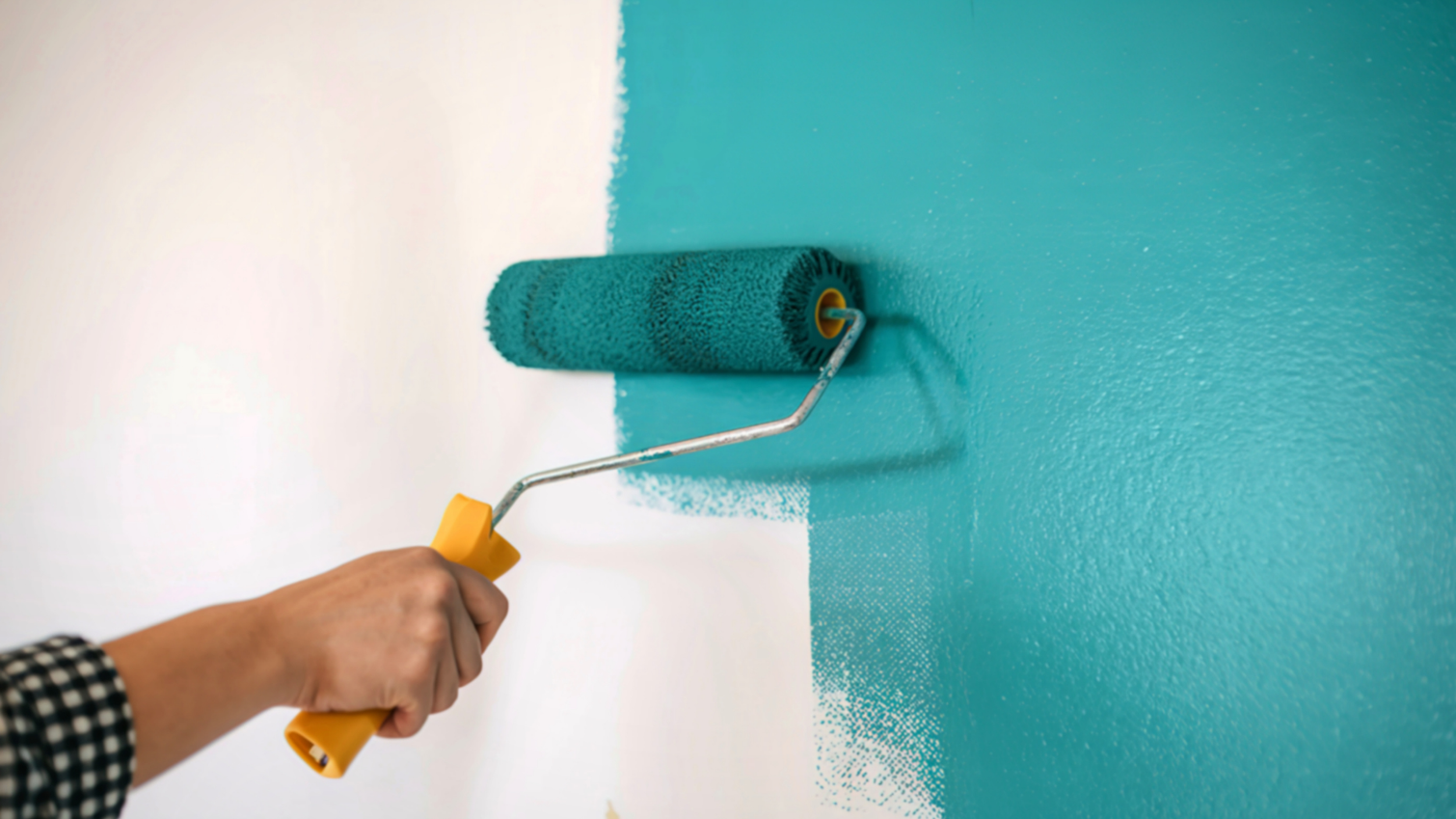 A hand holds a yellow-handled paint roller, applying bright teal paint to a previously white wall.