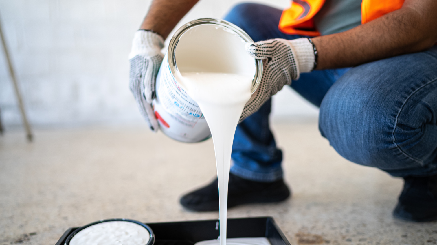 A worker in safety gear pours white paint from a metal can into a black tray on a concrete floor.