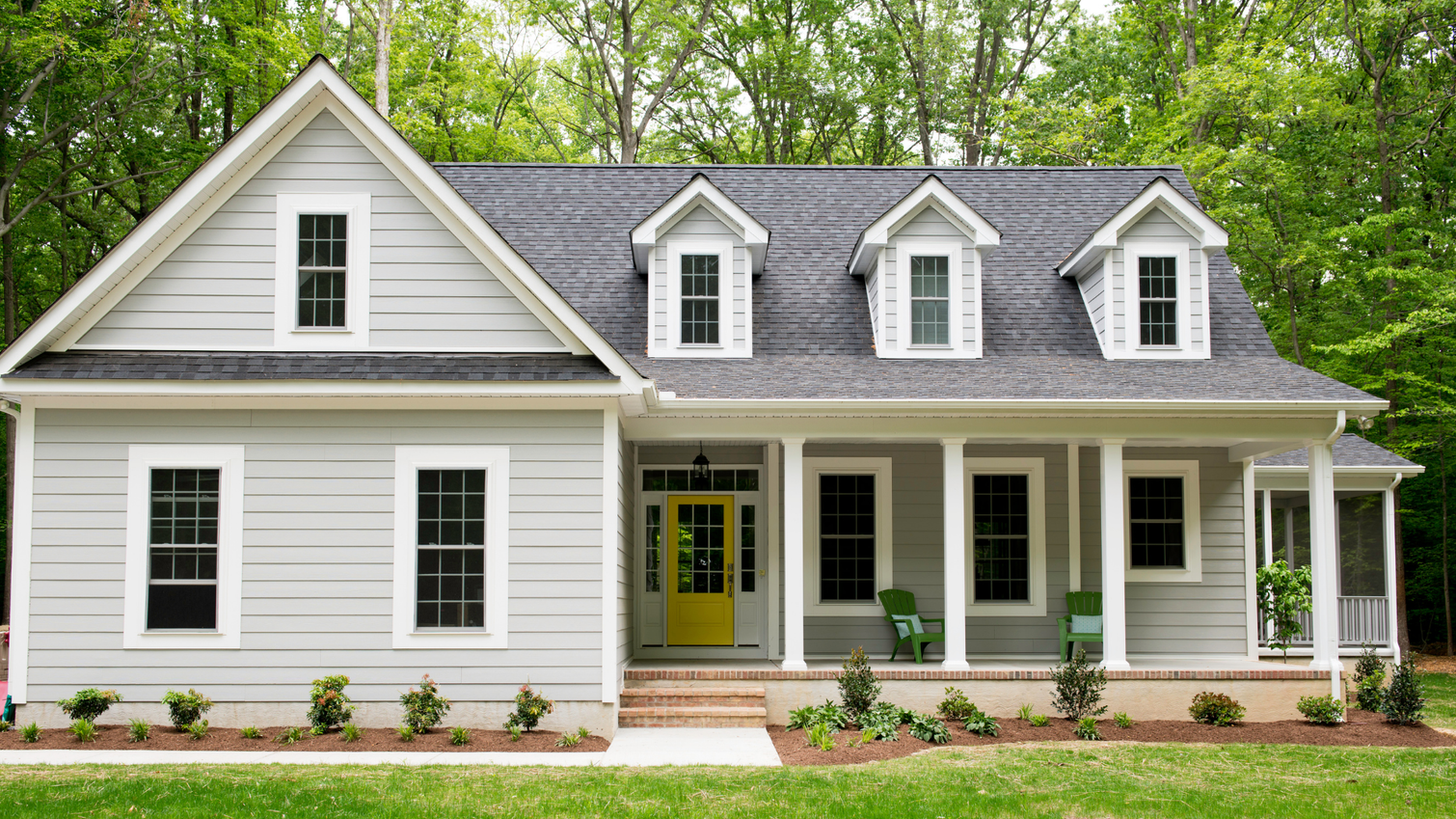 Light gray single-story house with a yellow front door, three dormer windows, and a covered porch in a wooded setting.