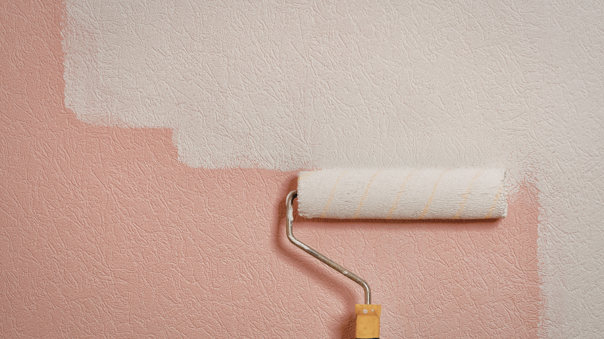 A paint roller applies a coat of white paint over a textured, pale pink wall.