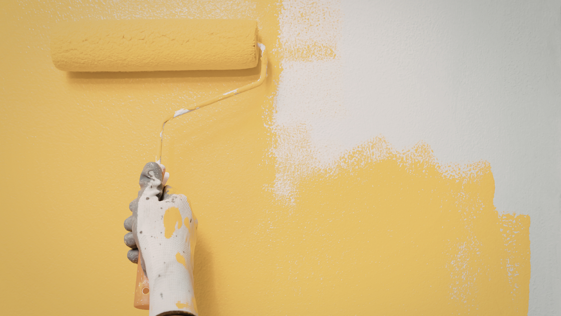 A gloved hand uses a paint roller to apply bright yellow paint over a white wall.
