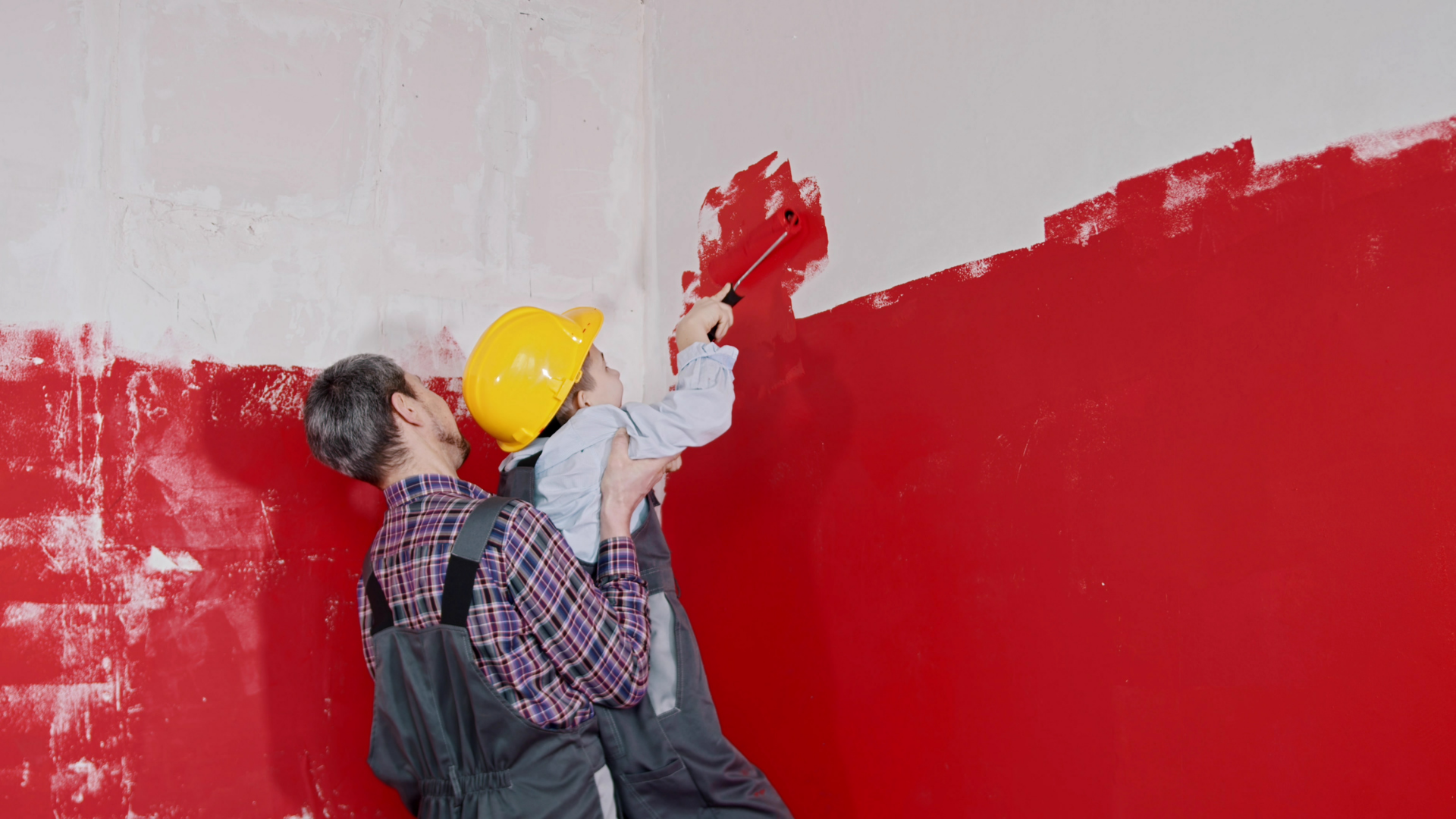 An adult in work clothes lifts a child wearing a yellow hard hat to help them paint a wall red with a small roller.