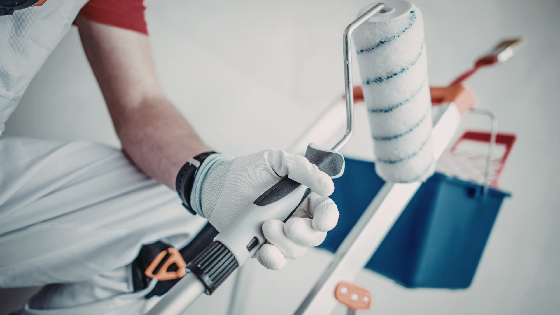 A gloved hand holds a paint roller handle attached to an extension pole, with a paint tray and bucket in the background.