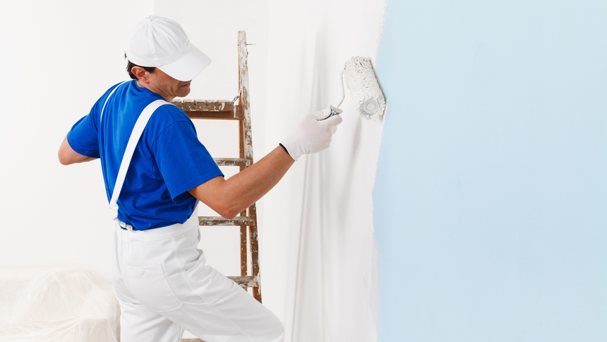 A painter in blue shirt and white overalls uses a roller to paint a wall light blue.