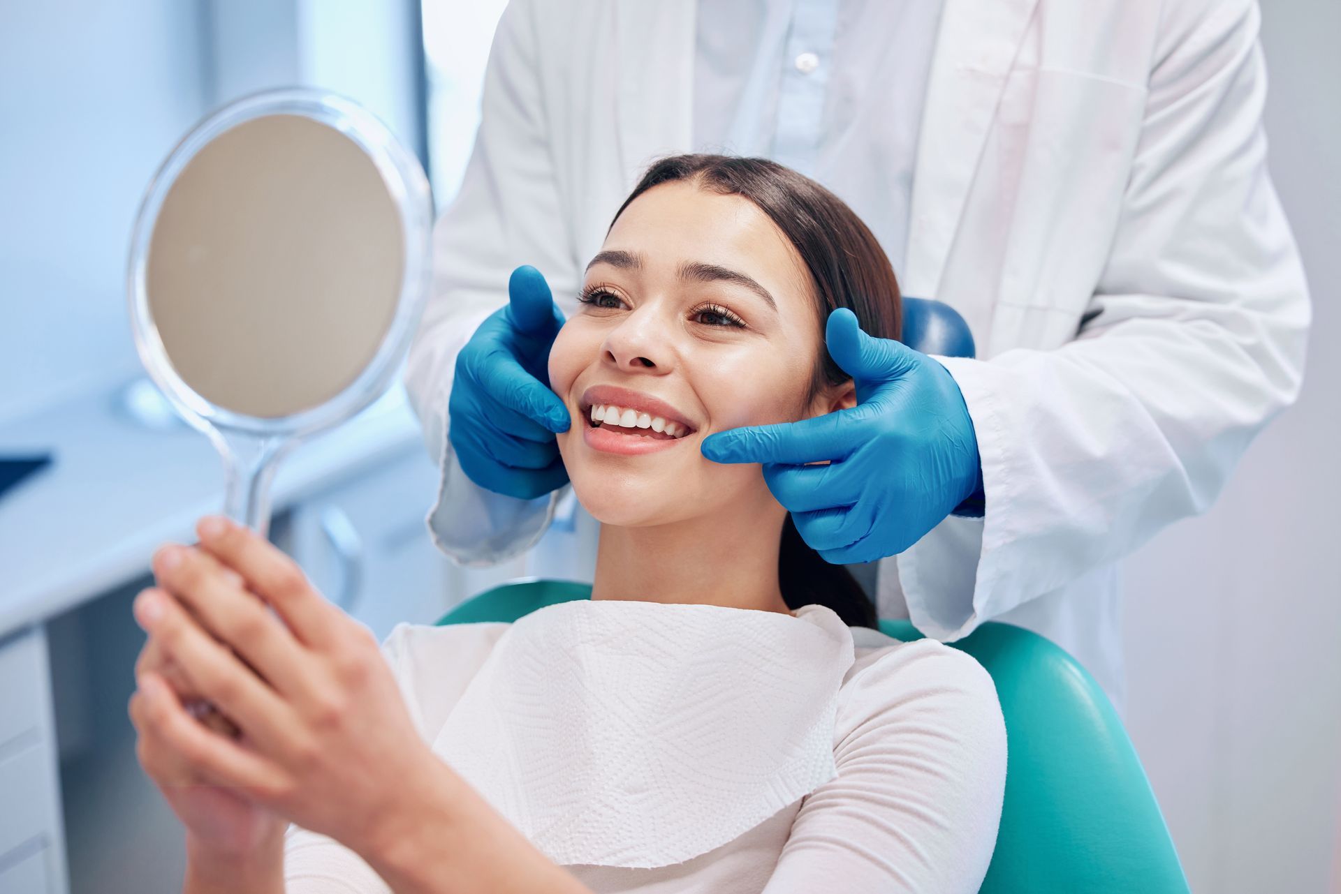 Dental patient holding mirror while clinician examines teeth with gloved hands.