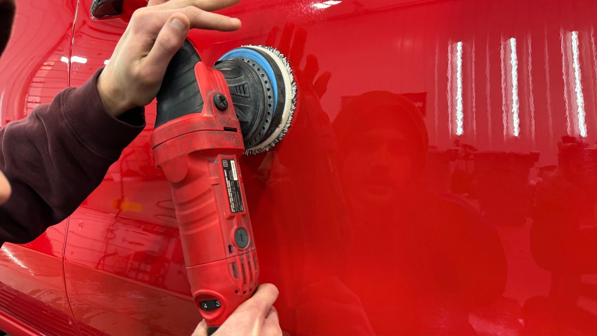 A person using a red dual-action polisher to buff a glossy red car surface in a bright workshop.