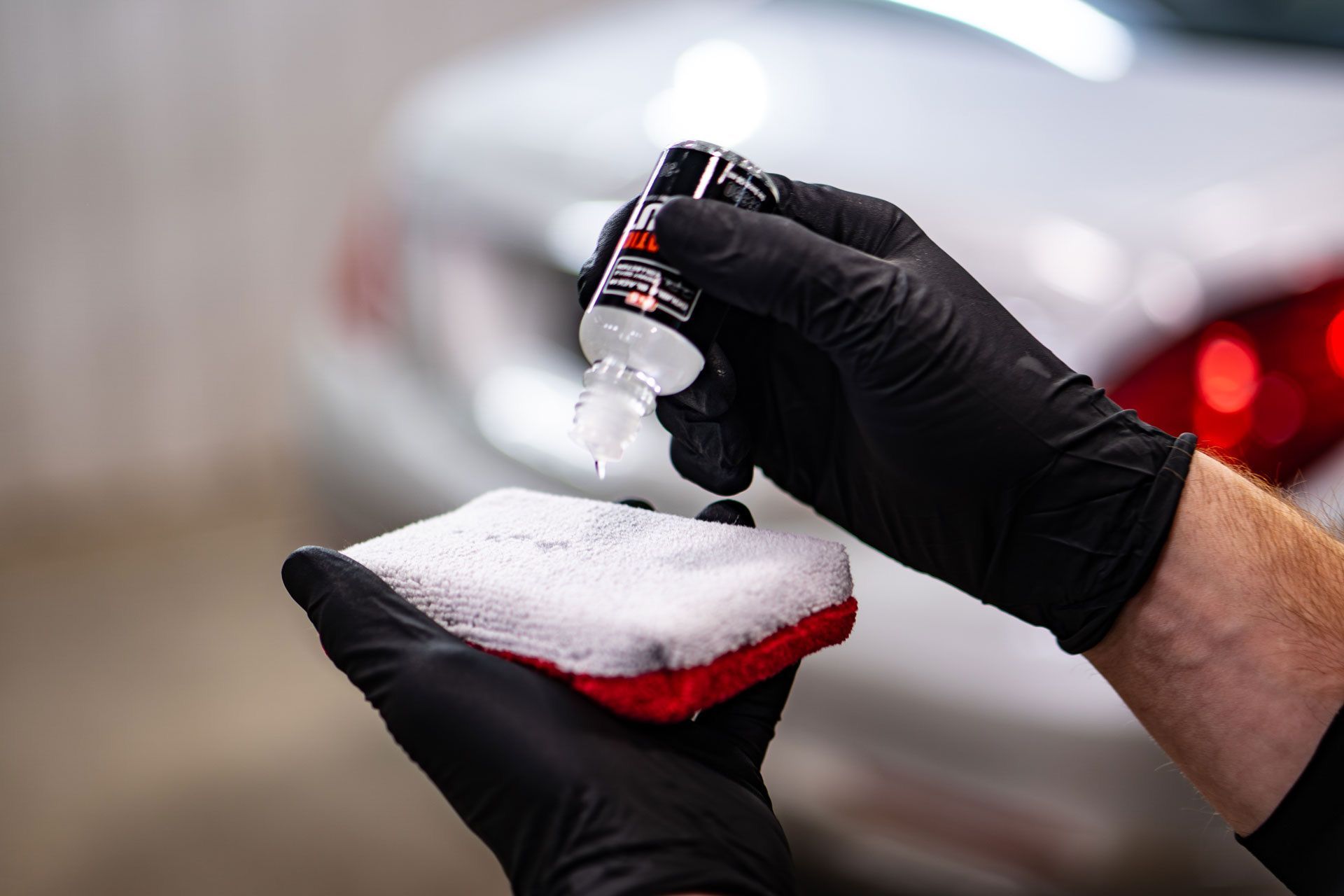 Hands in black gloves applying a liquid sealant from a small bottle onto a white applicator sponge for car detailing.