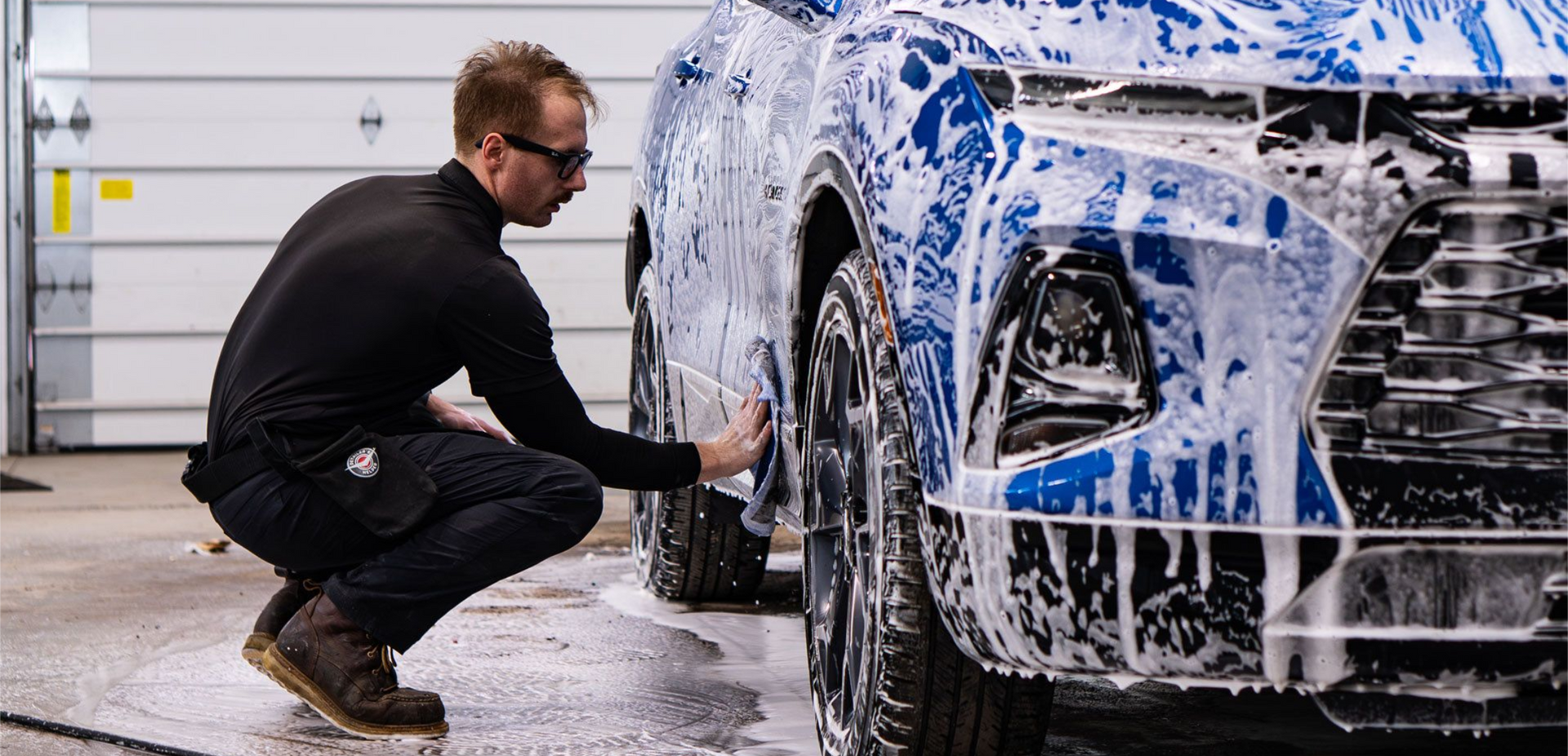 A person in a black turtleneck and work pants crouches to hand-wash a blue-foamed SUV in a brightly lit car wash bay.