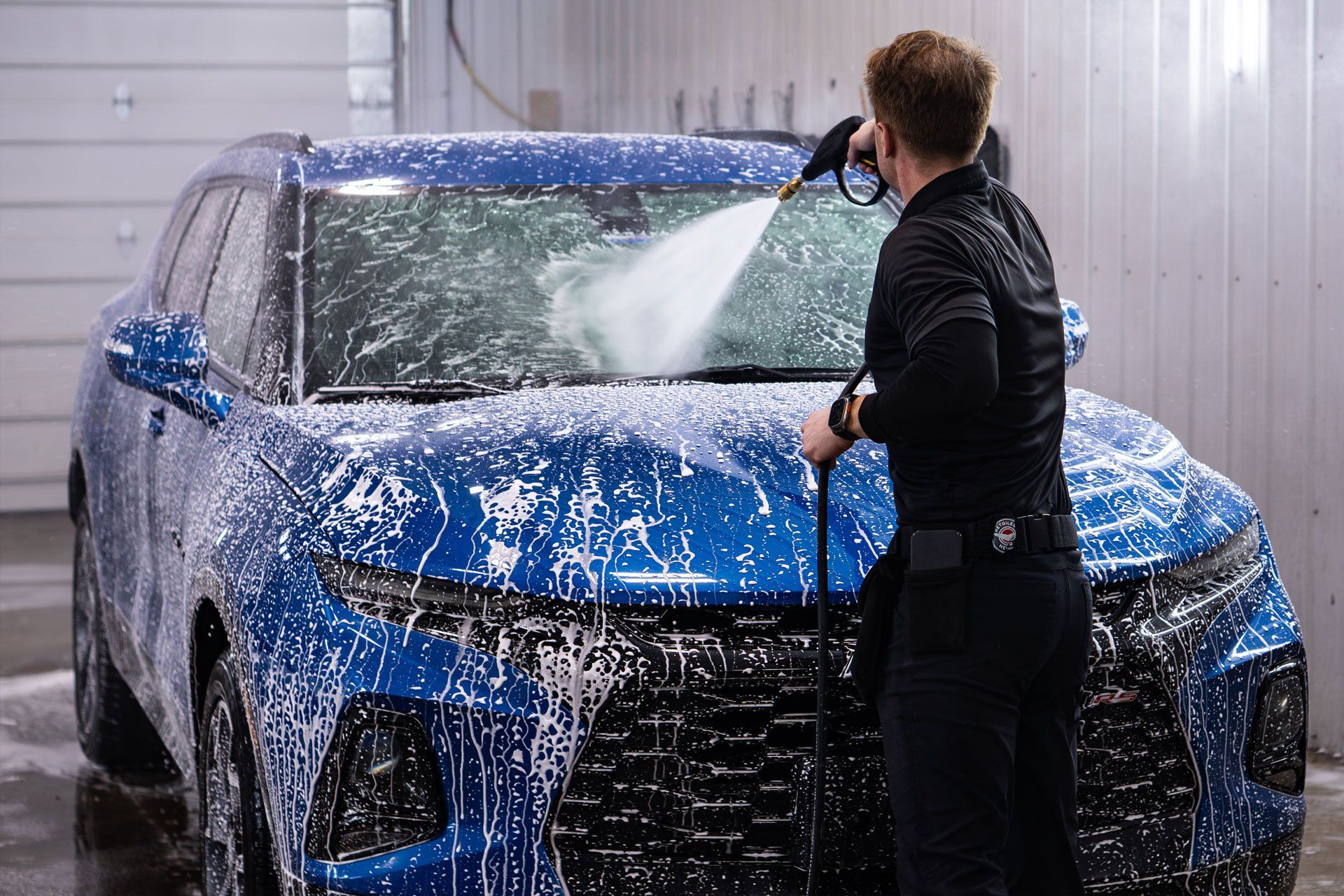 A person in a black uniform sprays foam onto a blue SUV in a car wash bay.