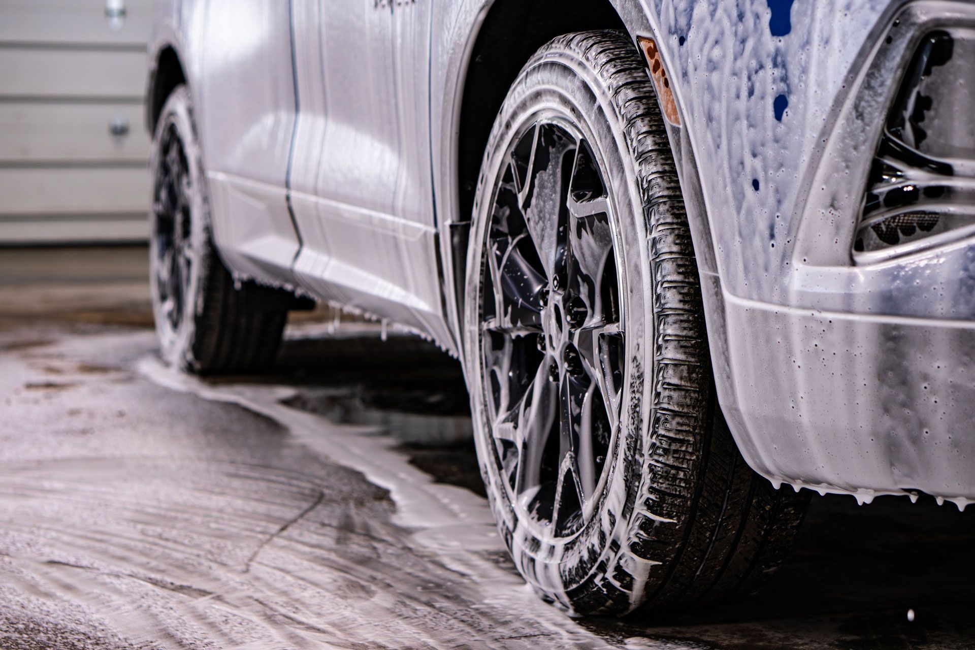 A car wheel and side panel covered in white cleaning foam during a car wash in a garage.