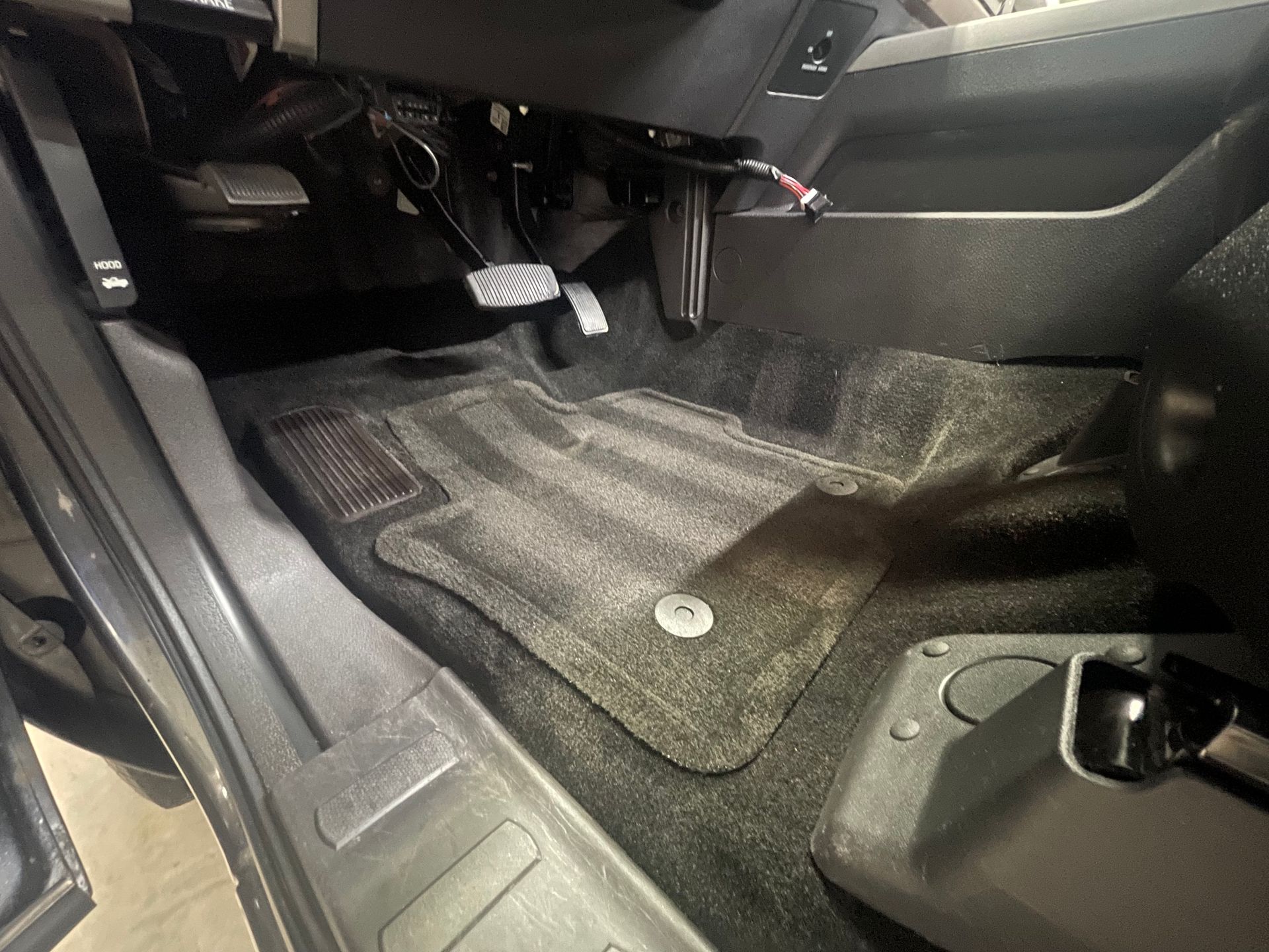 A grey car interior showing the driver-side footwell, pedals, and a secured grey floor mat.
