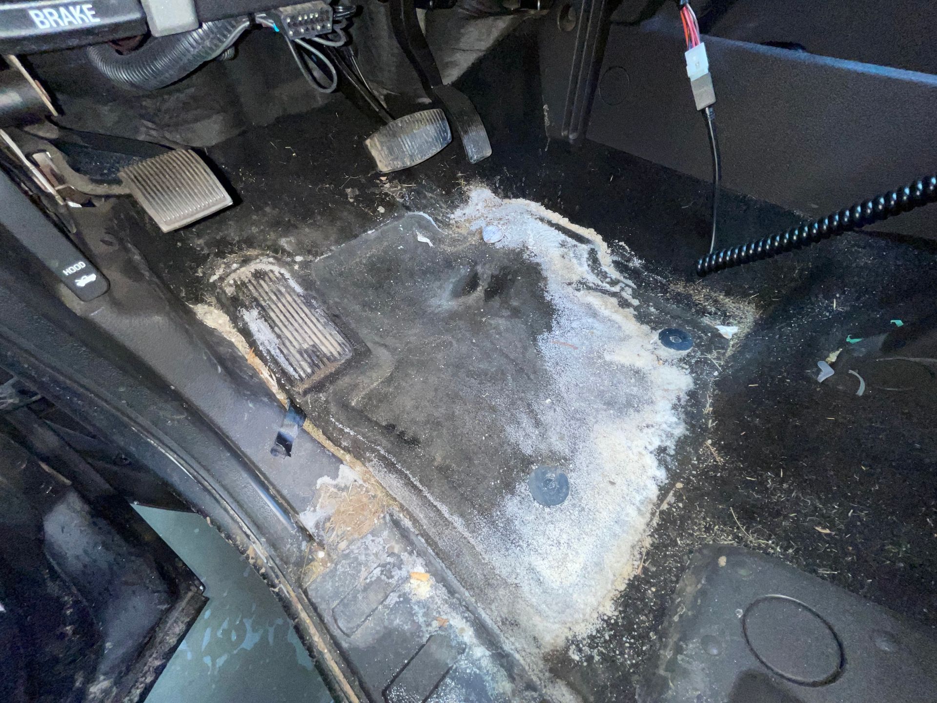 A dirty car interior floor showing significant salt stains, debris, and wear on the carpet near the pedals.