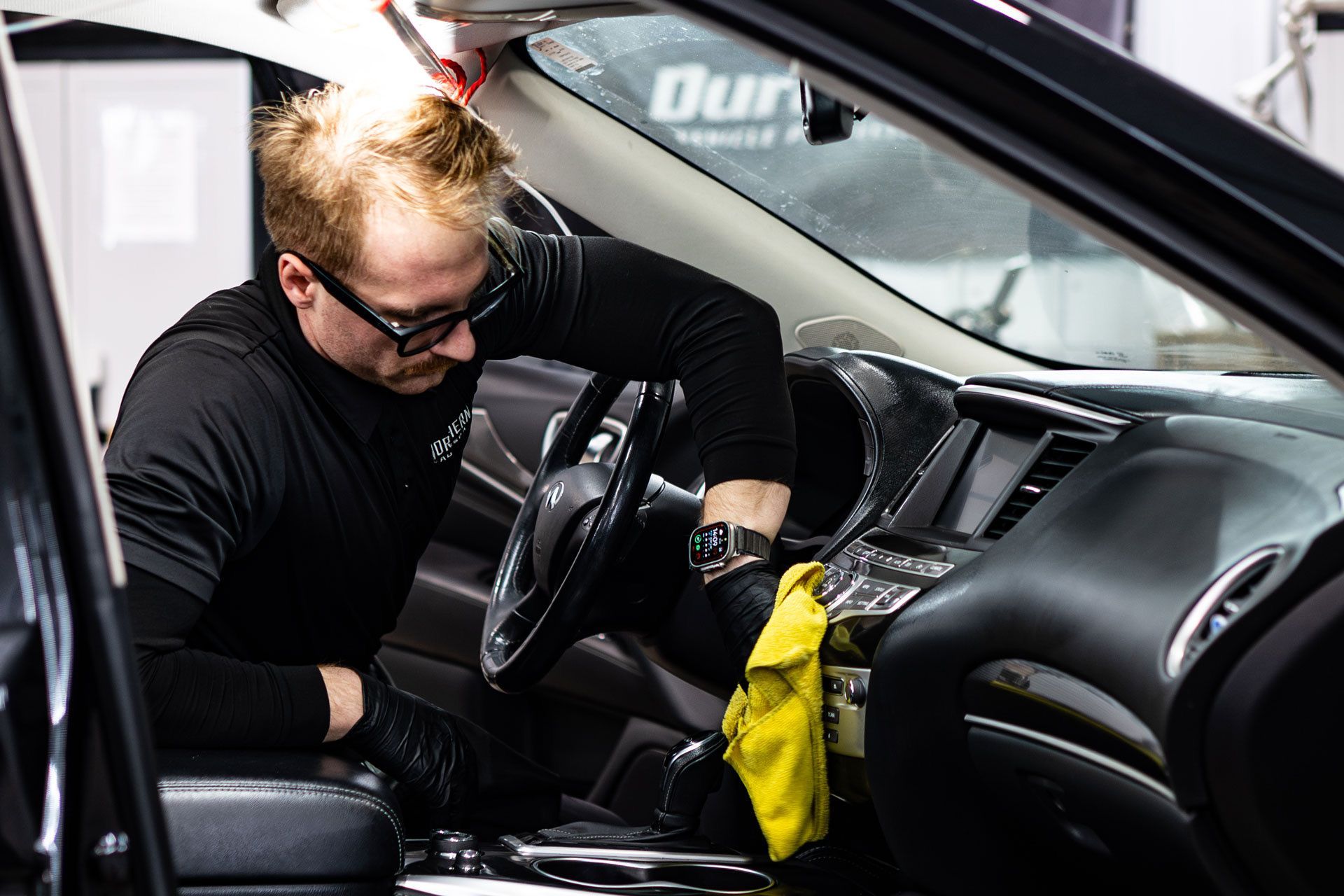 A person wearing black gloves cleans the interior dashboard of a car with a yellow microfiber cloth.