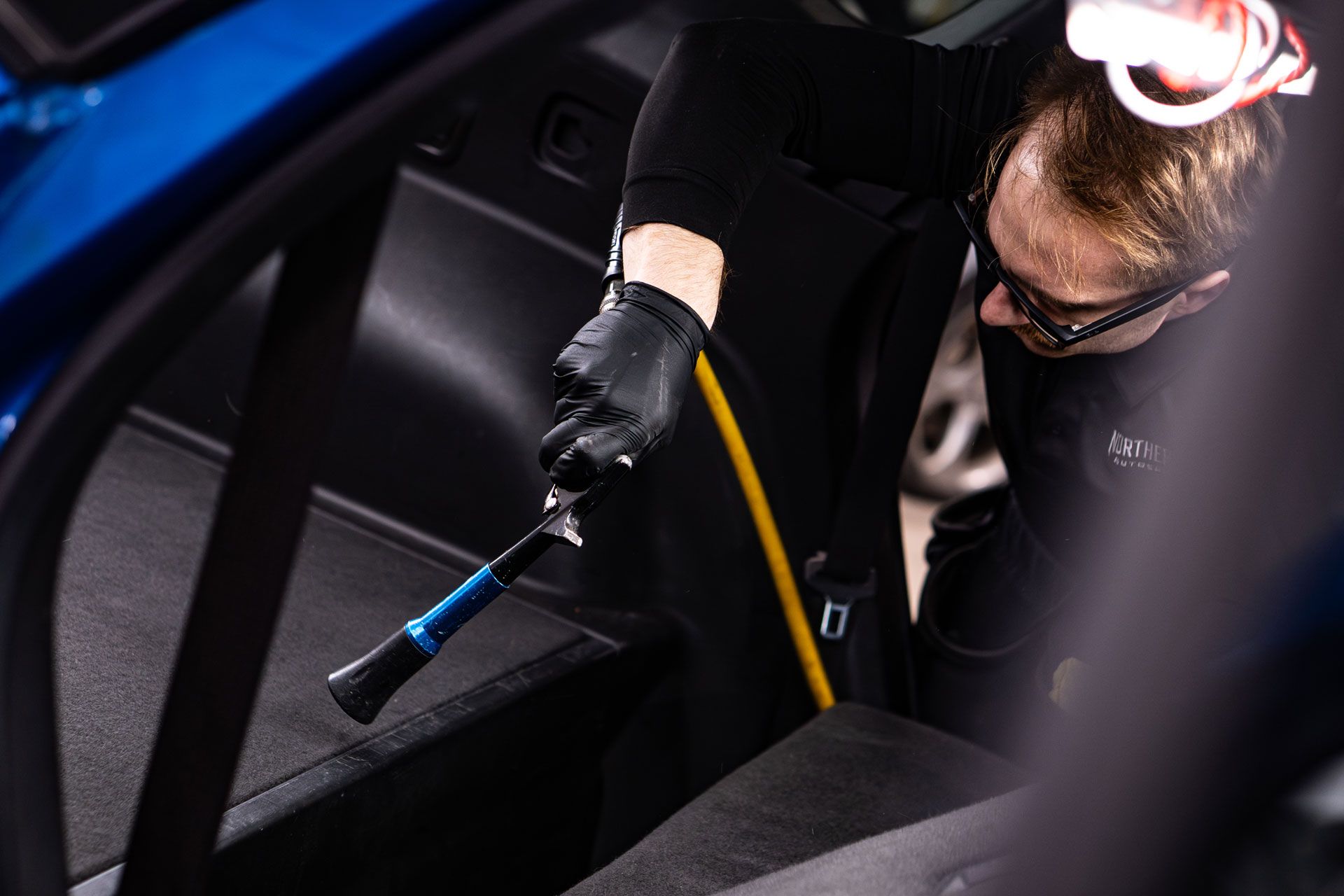A technician wearing black gloves uses a compressed air cleaning tool to detail the interior carpet of a blue vehicle.