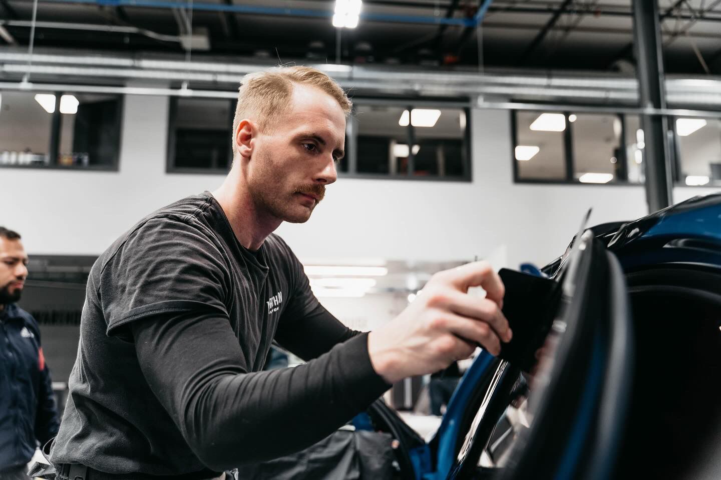 A worker in a dark t-shirt carefully applies a film or trim to the door frame of a blue vehicle in a workshop.