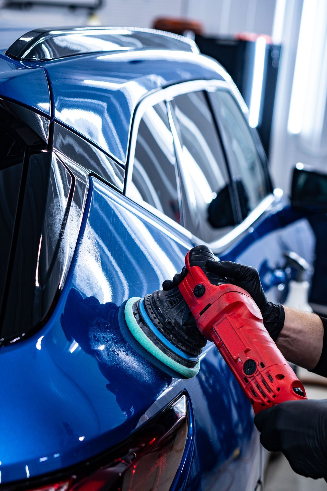 A person in black gloves uses a red power buffer with a blue pad to polish the glossy blue exterior of a vehicle.