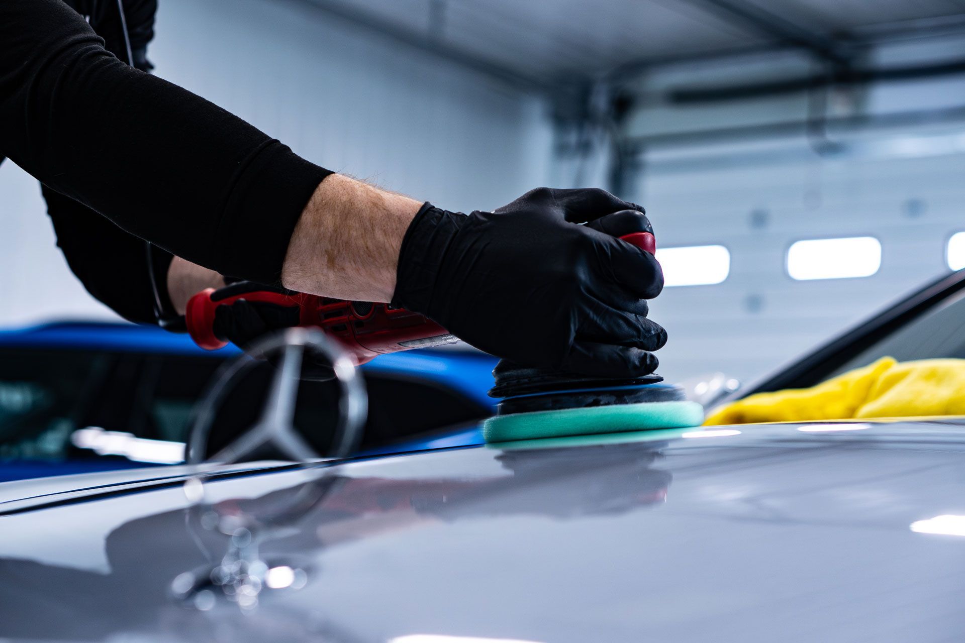 A worker in black gloves uses a power polisher with a green foam pad on the hood of a silver Mercedes-Benz in a garage.