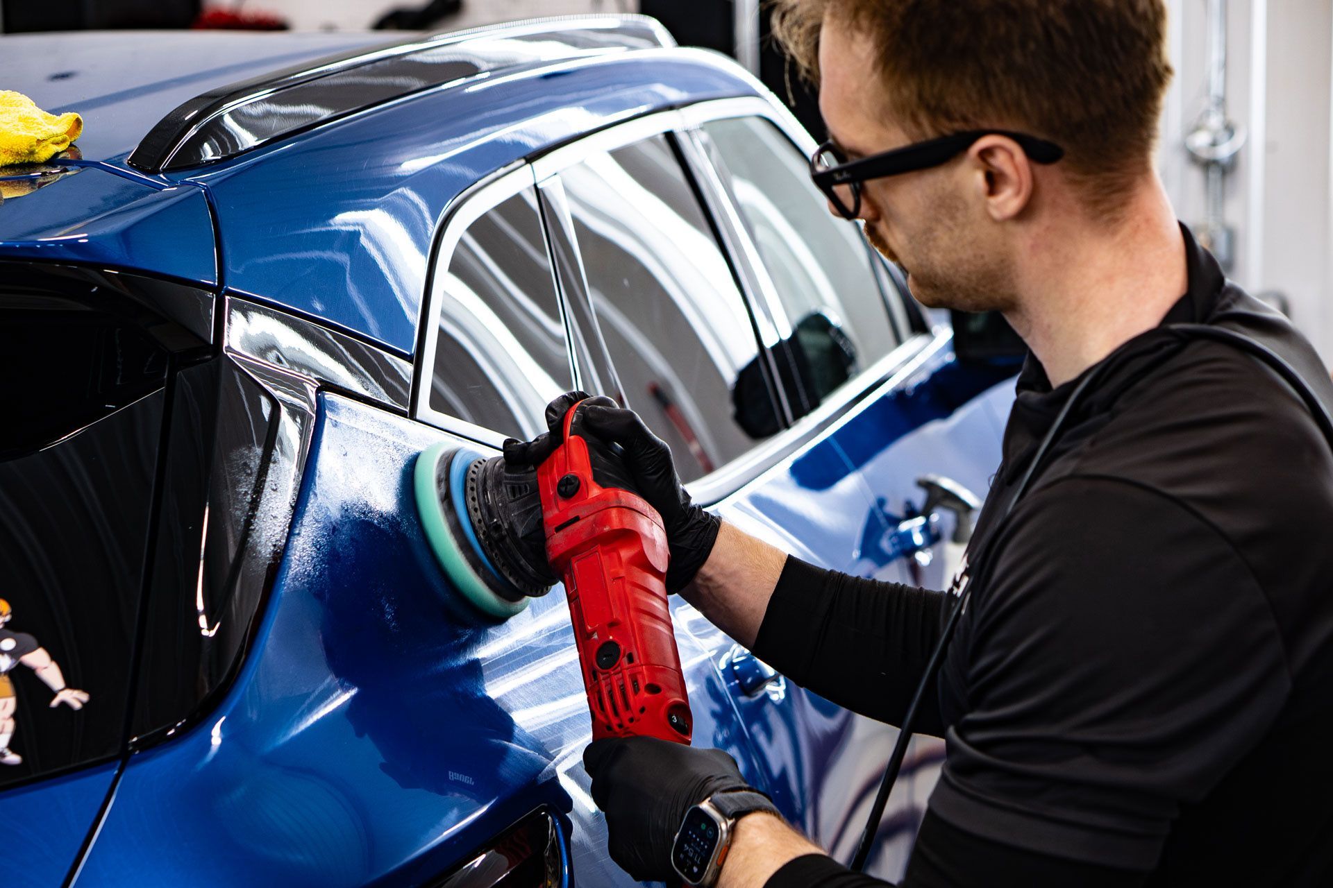A person wearing glasses and black gloves uses a red power polisher on the blue rear panel of a car.
